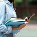 Student girl holding books and smartphone while walking in school campus background, education, back to school concept